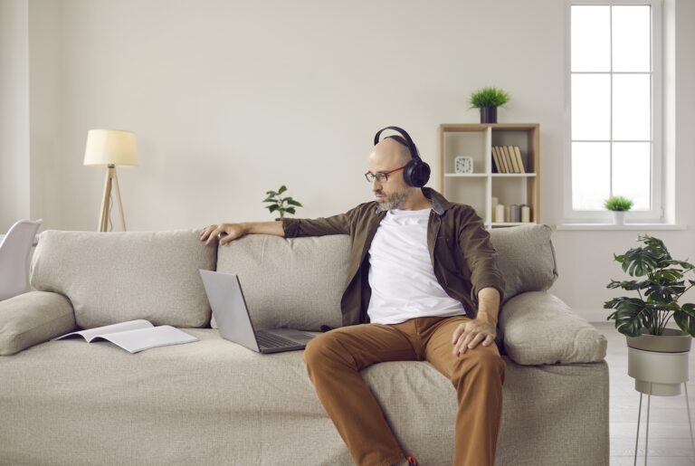 Mature man in headphones sitting on sofa at home and watching video on laptop computer
